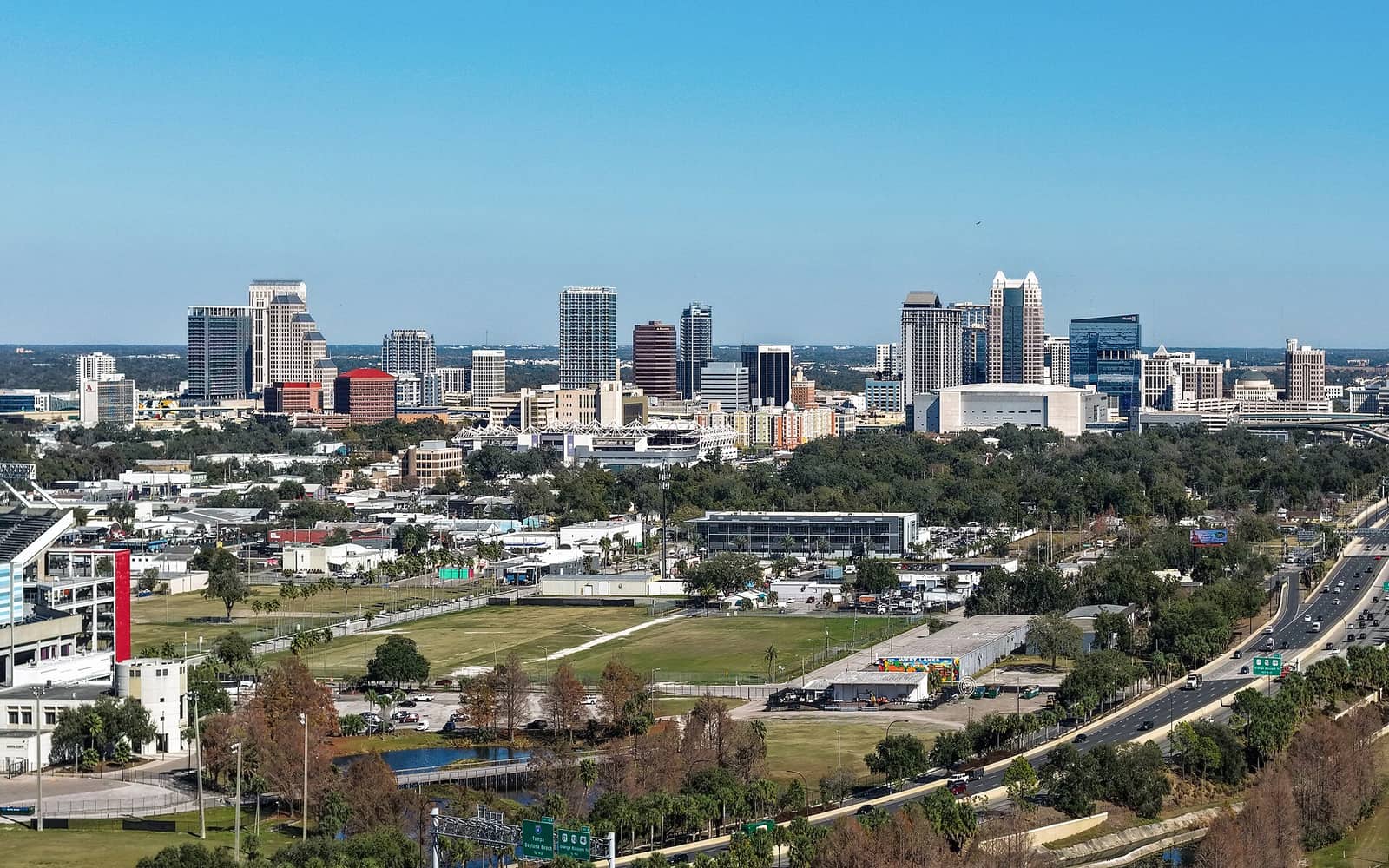 Elevated daytime view of a city skyline with open fields, trees, and a highway in front.
