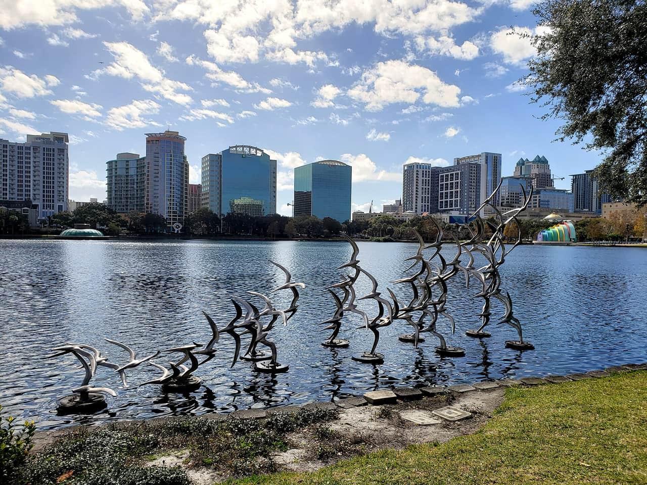 Metal bird sculptures stand in a sunlit lake with a downtown skyline of towers behind.