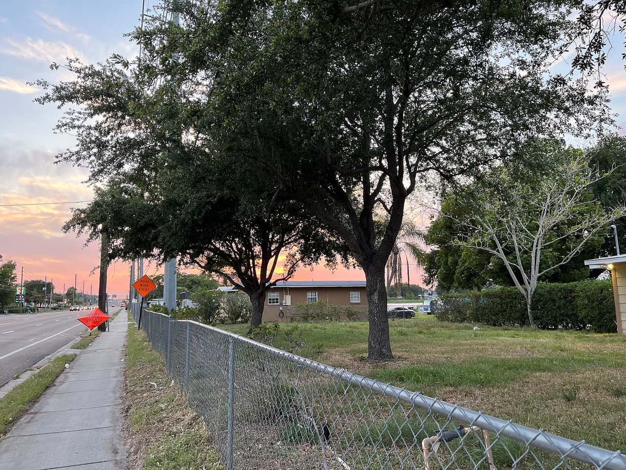 Tree-lined street with a chain-link fenced grassy lot and a low brick house under a sunset sky.