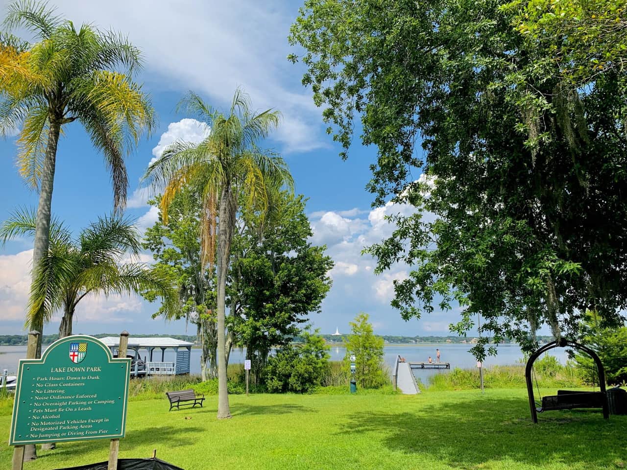 Grassy lakeside park with palm trees, benches, a park rules sign and a view across open water.