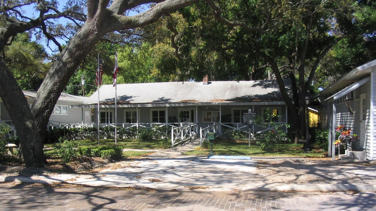 Historic low white wooden building with a porch, flagpoles and large oak trees shading the front lawn.