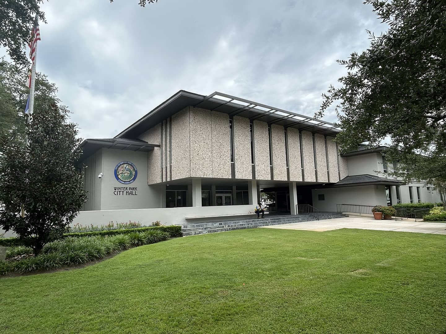 Modernist city hall with a flagpole and lawn under a cloudy sky, with a seal on the facade.