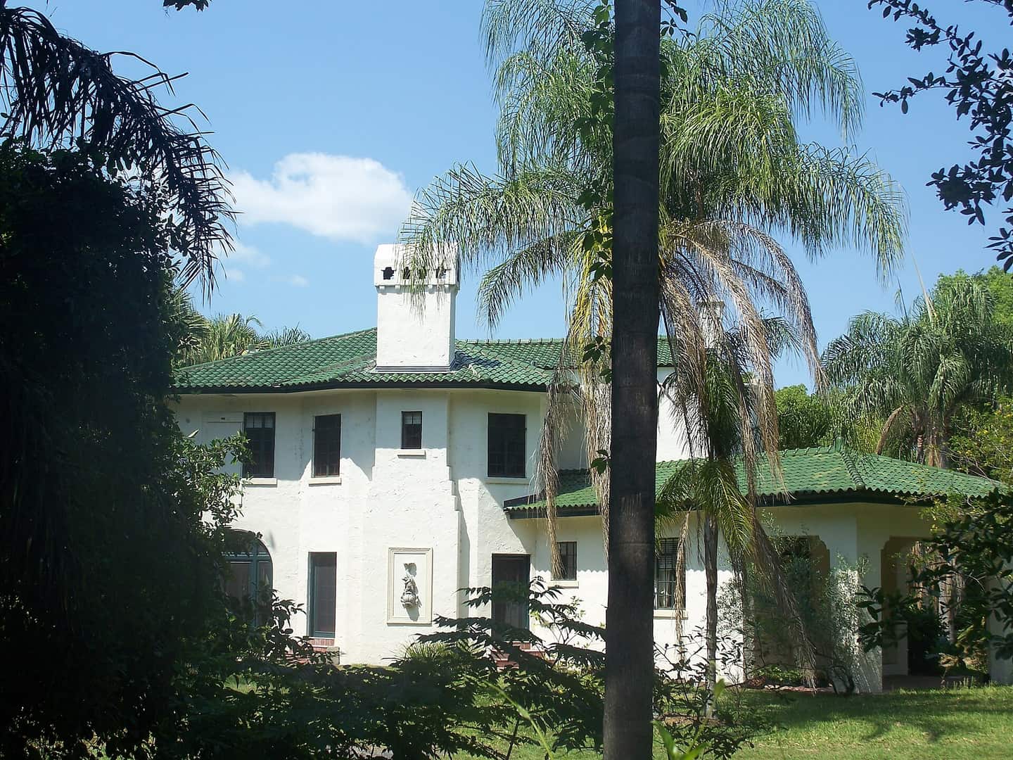 White stucco historic house with a green tile roof and tall palm trees under a partly cloudy sky.