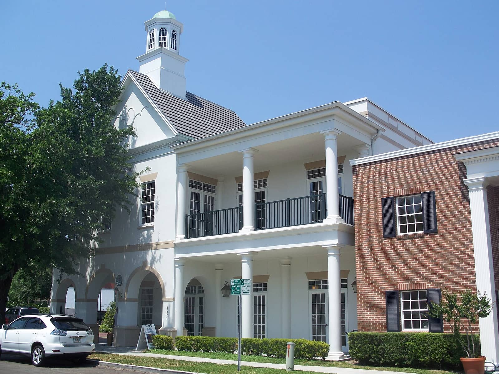 White colonial-style welcome center with columns and a balcony beside a red brick building under blue sky.