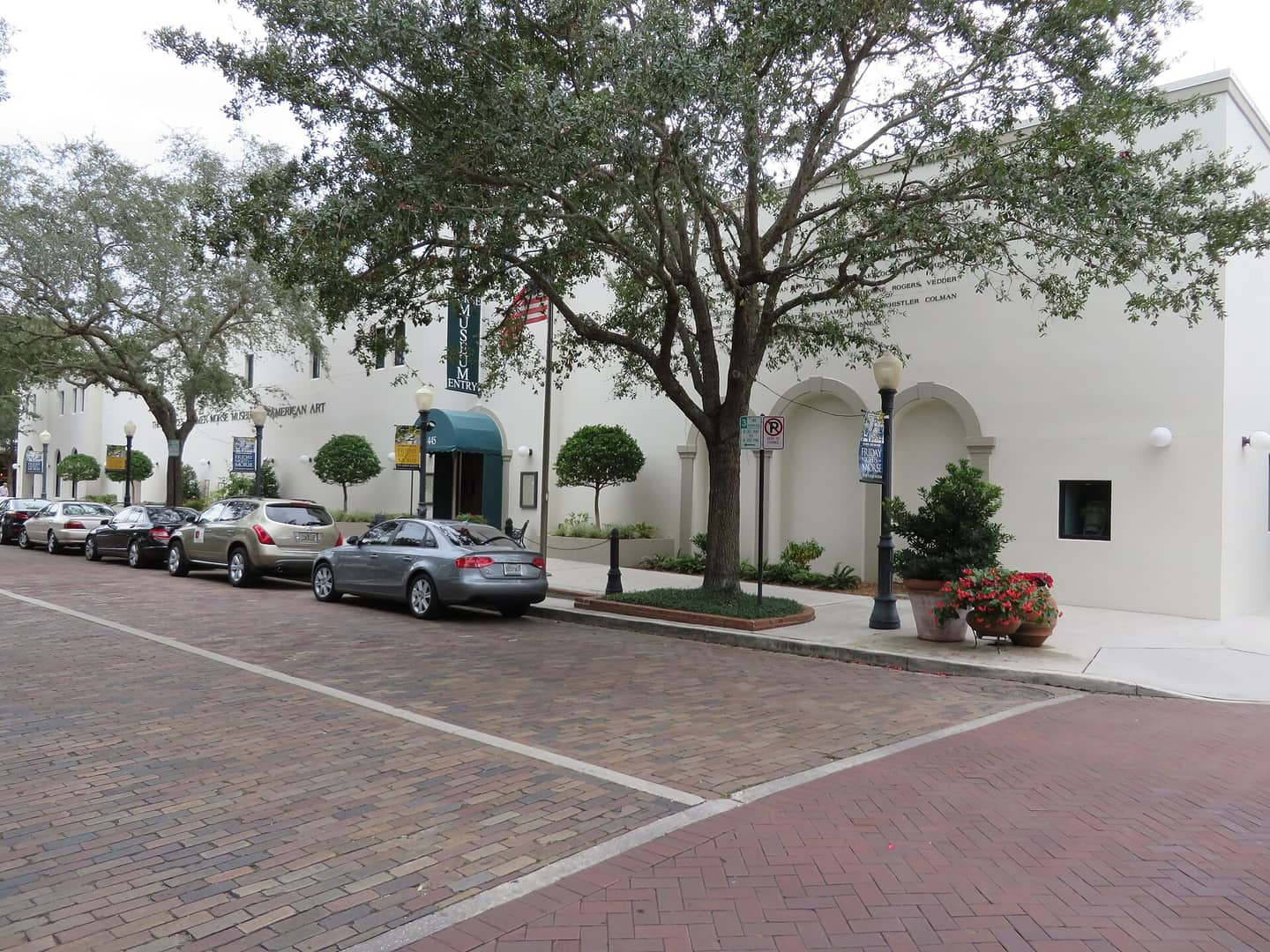 White stucco museum building with arched entrance beside a brick street and parked cars under trees.