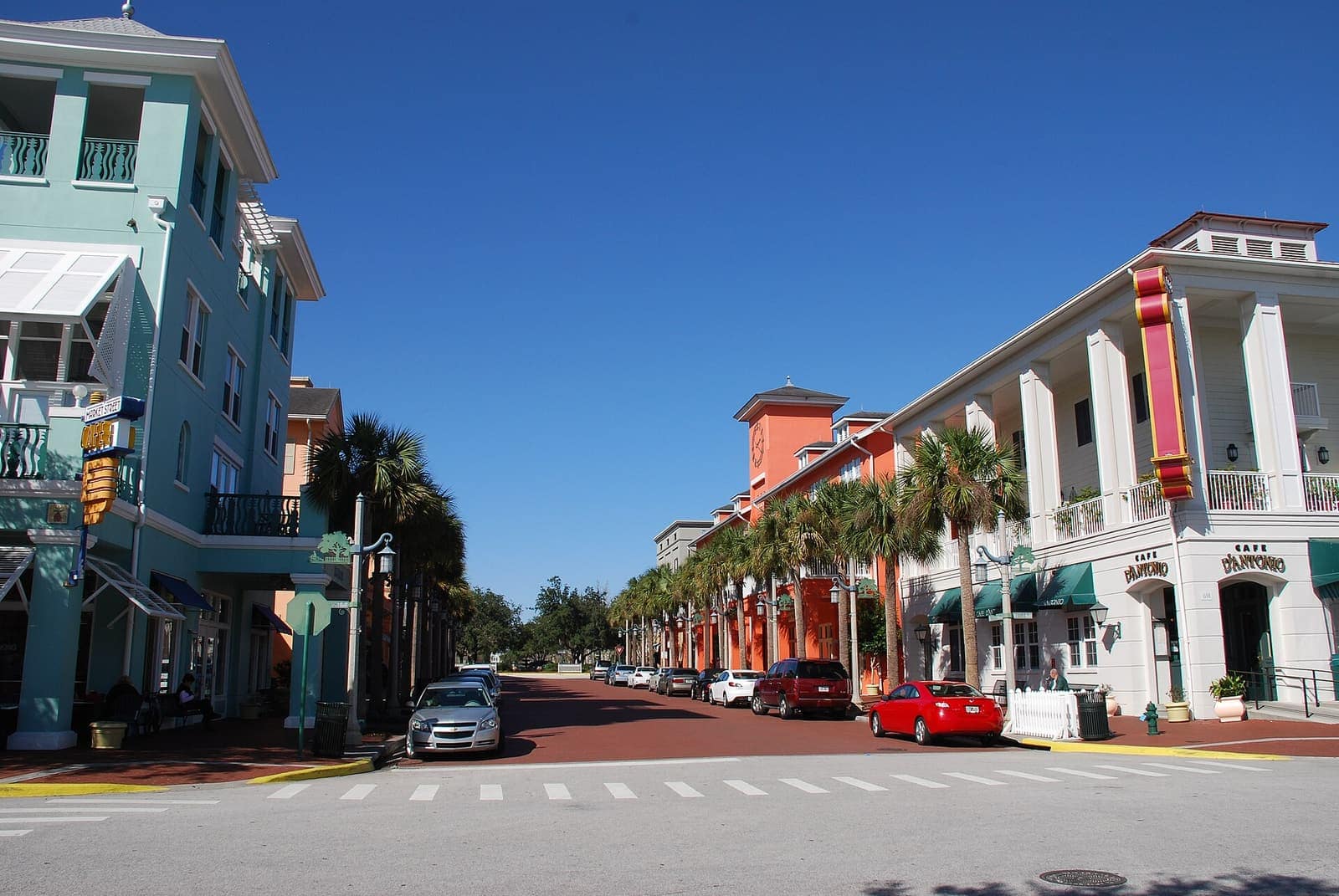 Wide downtown street lined with colourful two-storey buildings, palm trees, parked cars and a clear blue sky.