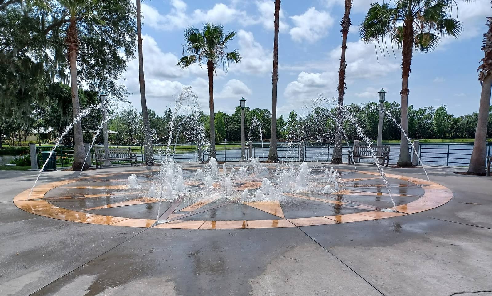 Circular fountain with water jets on a paved plaza, with palm trees and a lake behind it.
