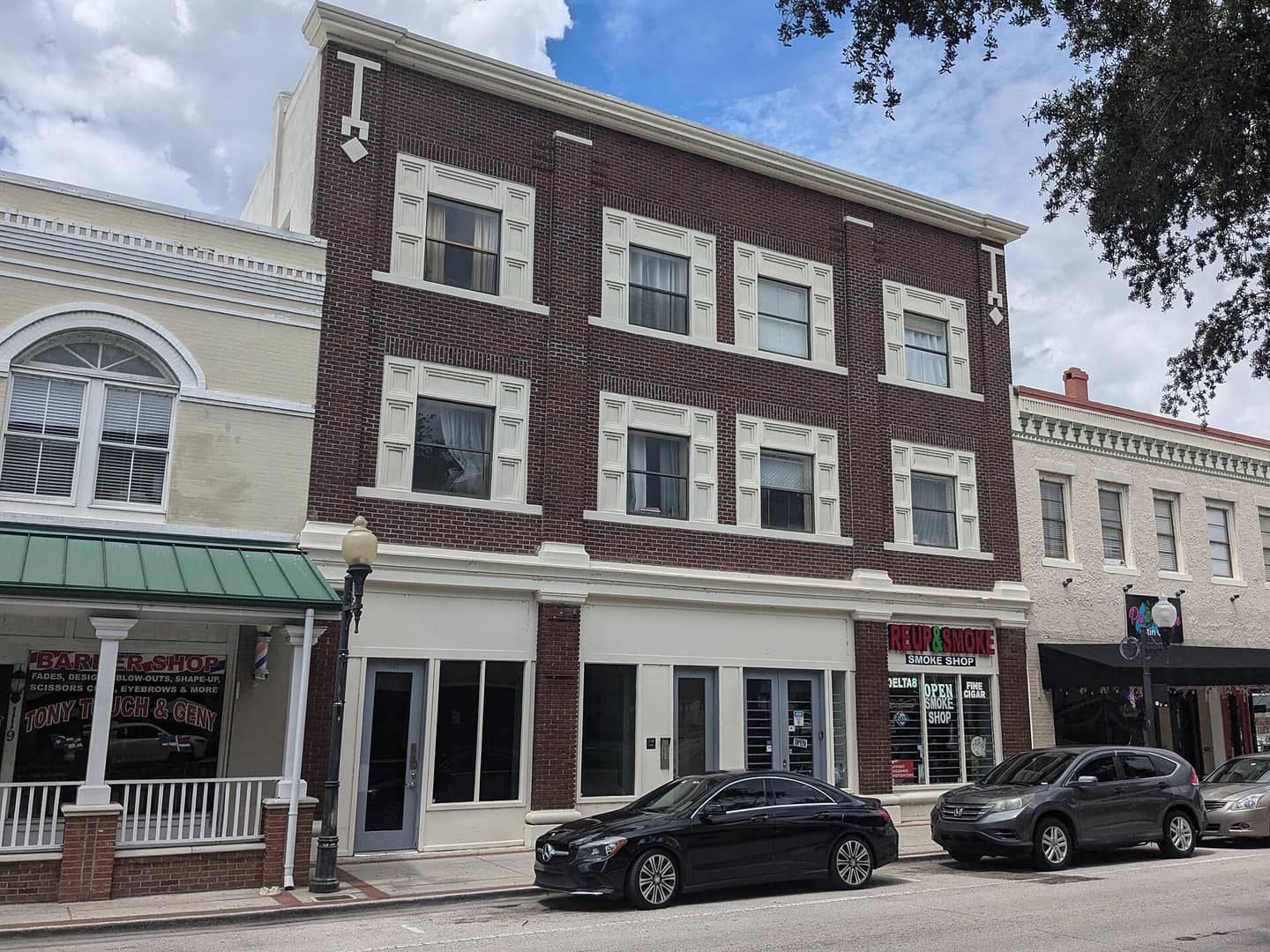 Three-storey red brick commercial building with white window trim beside a green-awning storefront and parked cars.