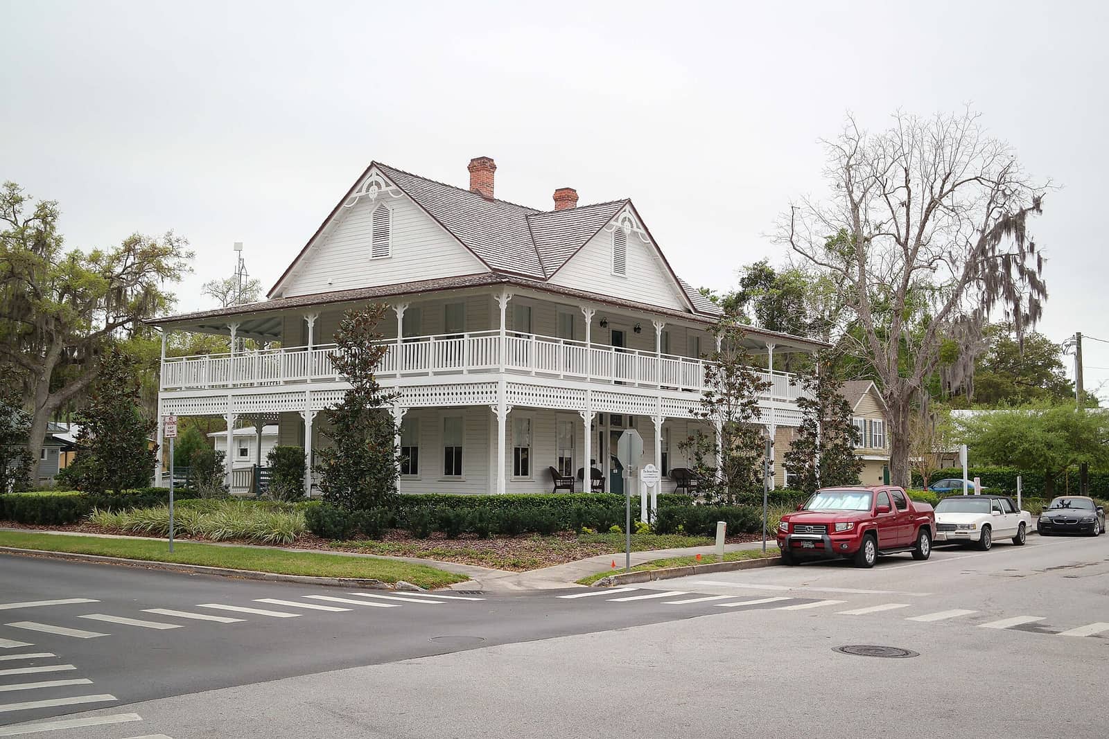 White two-story Victorian house with a wraparound second-floor porch, shaded by oak trees, beside a street with parked cars.