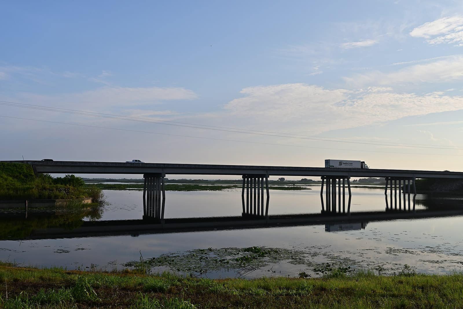 Highway bridge crossing a calm river at dusk, with a truck and cars on the deck and reflections in water.
