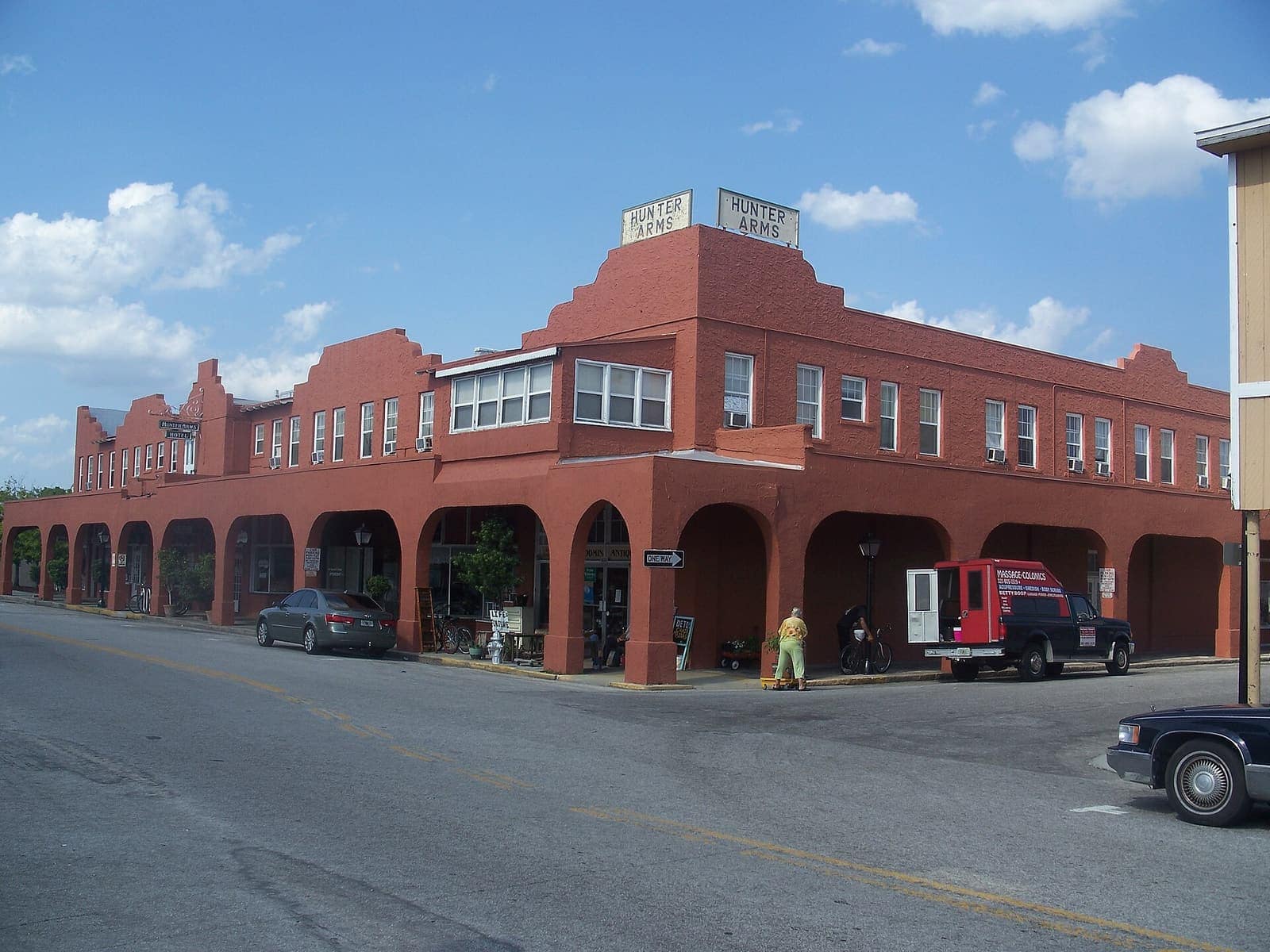 Red brick arcaded building with a rooftop sign and parked vehicles along a sunny street.