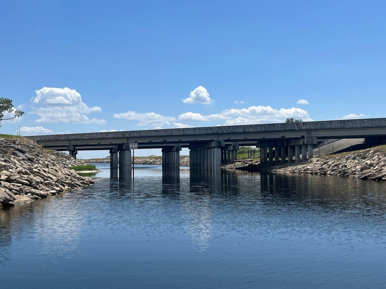 Concrete road bridge spanning a calm canal with rock embankments under a bright blue sky.