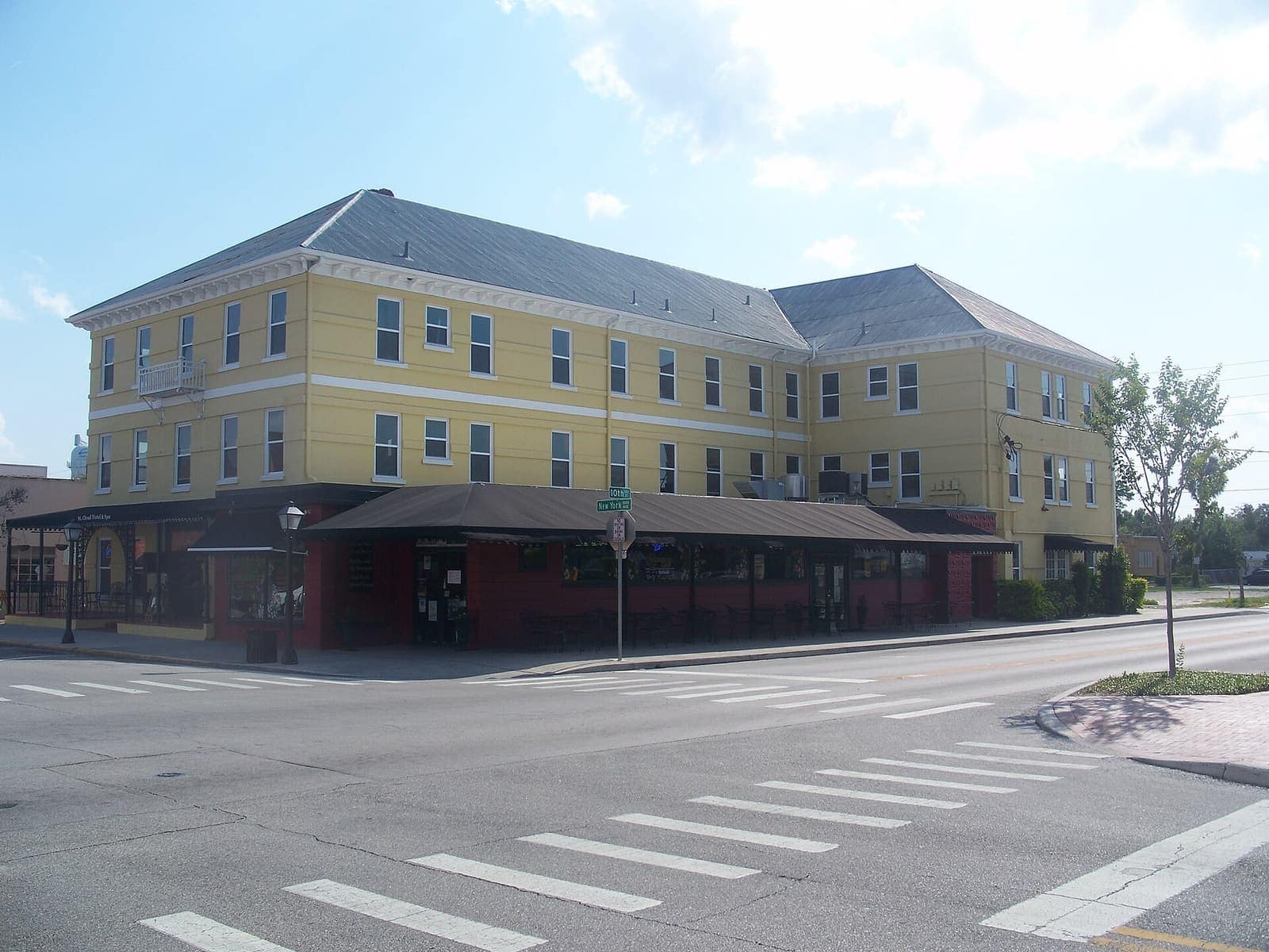 Yellow three-story historic hotel with a long red-trimmed arcade along a sunny street corner.