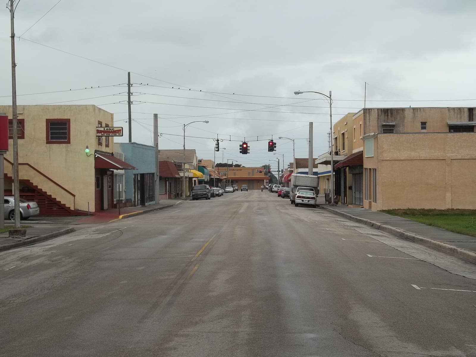 Overcast daytime view down a small-town street with low storefronts, traffic signals, and parked cars on both sides.