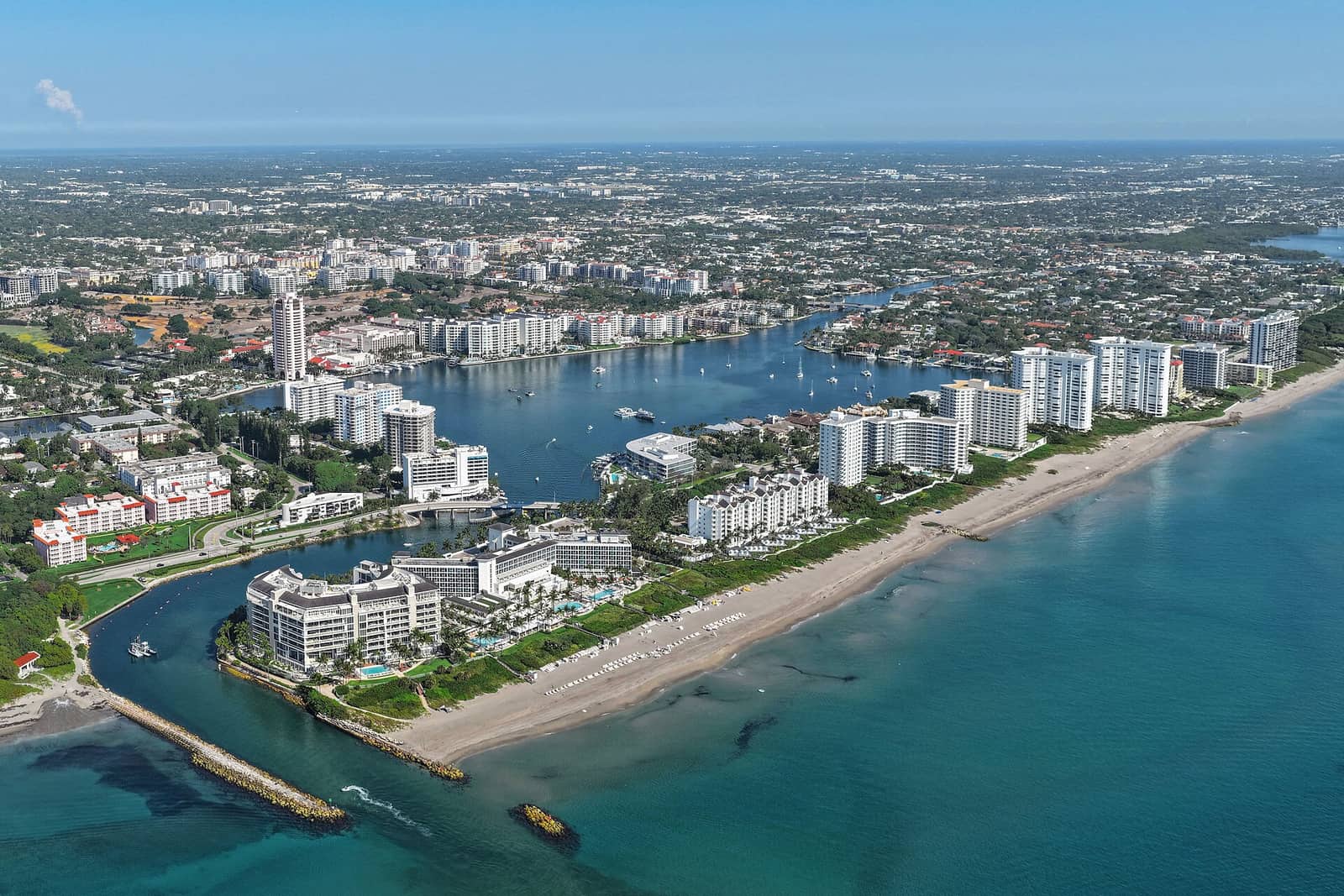 Aerial view of a coastal inlet, high-rise towers, a lake, and a long sandy beachfront under clear sky.