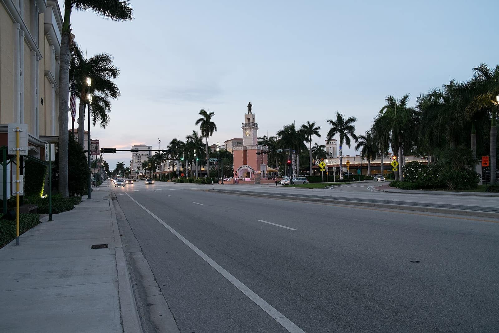 Wide boulevard at dusk leads toward a pink clock tower with a statue, flanked by tall palm trees.
