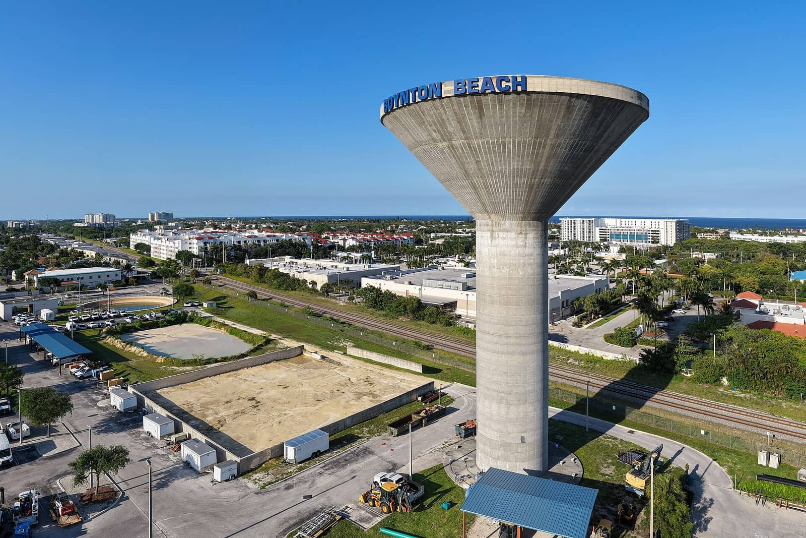 Aerial view of a concrete water tower bearing Boynton Beach lettering, with buildings, a rail line and the ocean beyond.