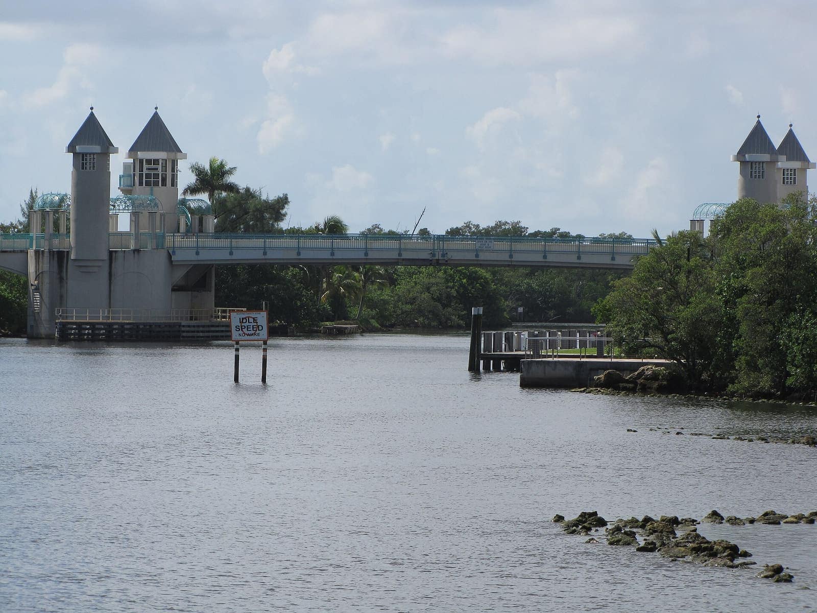 A gray drawbridge with two corner towers spans a calm waterway under a partly cloudy sky with green trees.