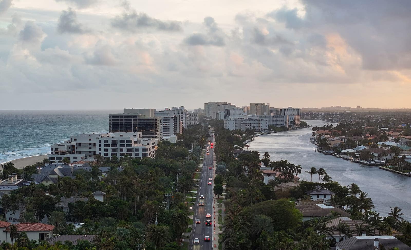 Aerial dusk view of beachfront high-rises, a palm-lined road with cars, and a waterway.