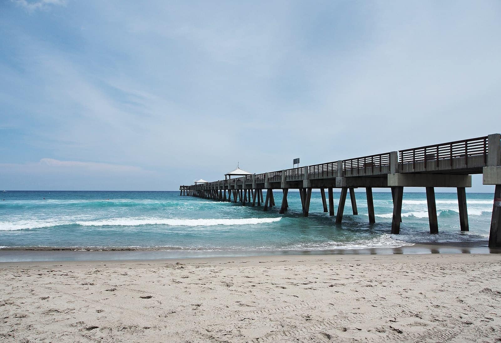 A long wooden pier extends into rolling ocean waves from a pale sandy beach under a cloudy sky.
