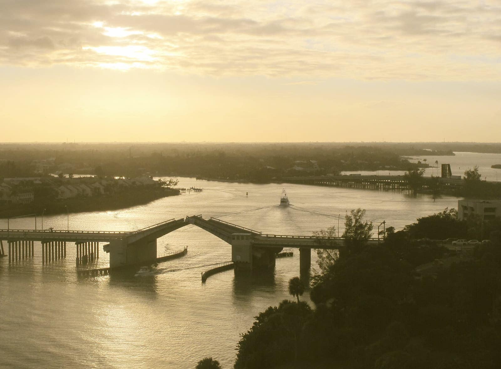 A drawbridge partly raised over a waterway at sunset, with a boat passing and palm-lined shoreline in the distance.