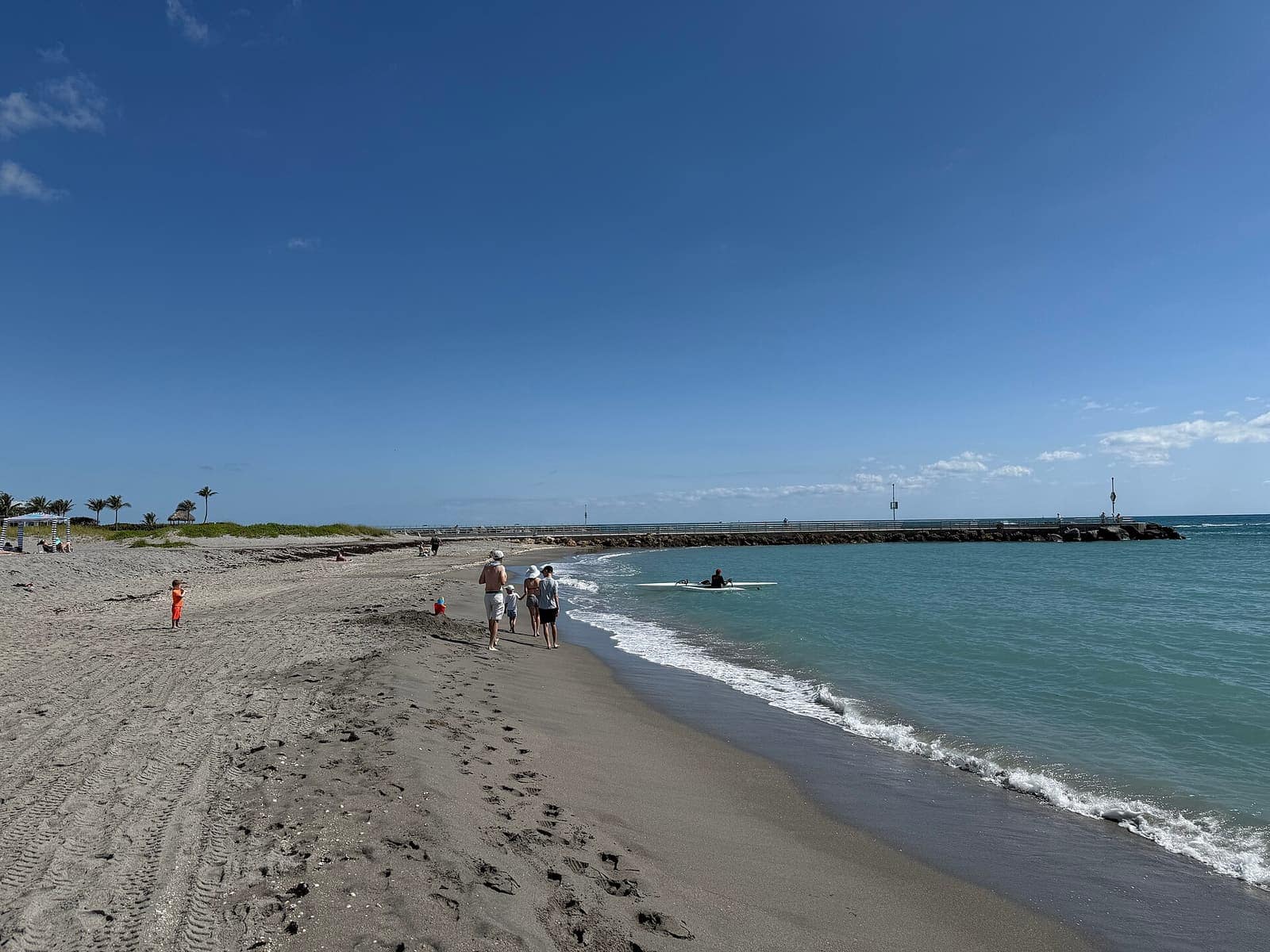 Beachgoers walk on sand beside turquoise surf, with a rock jetty stretching into the sea.