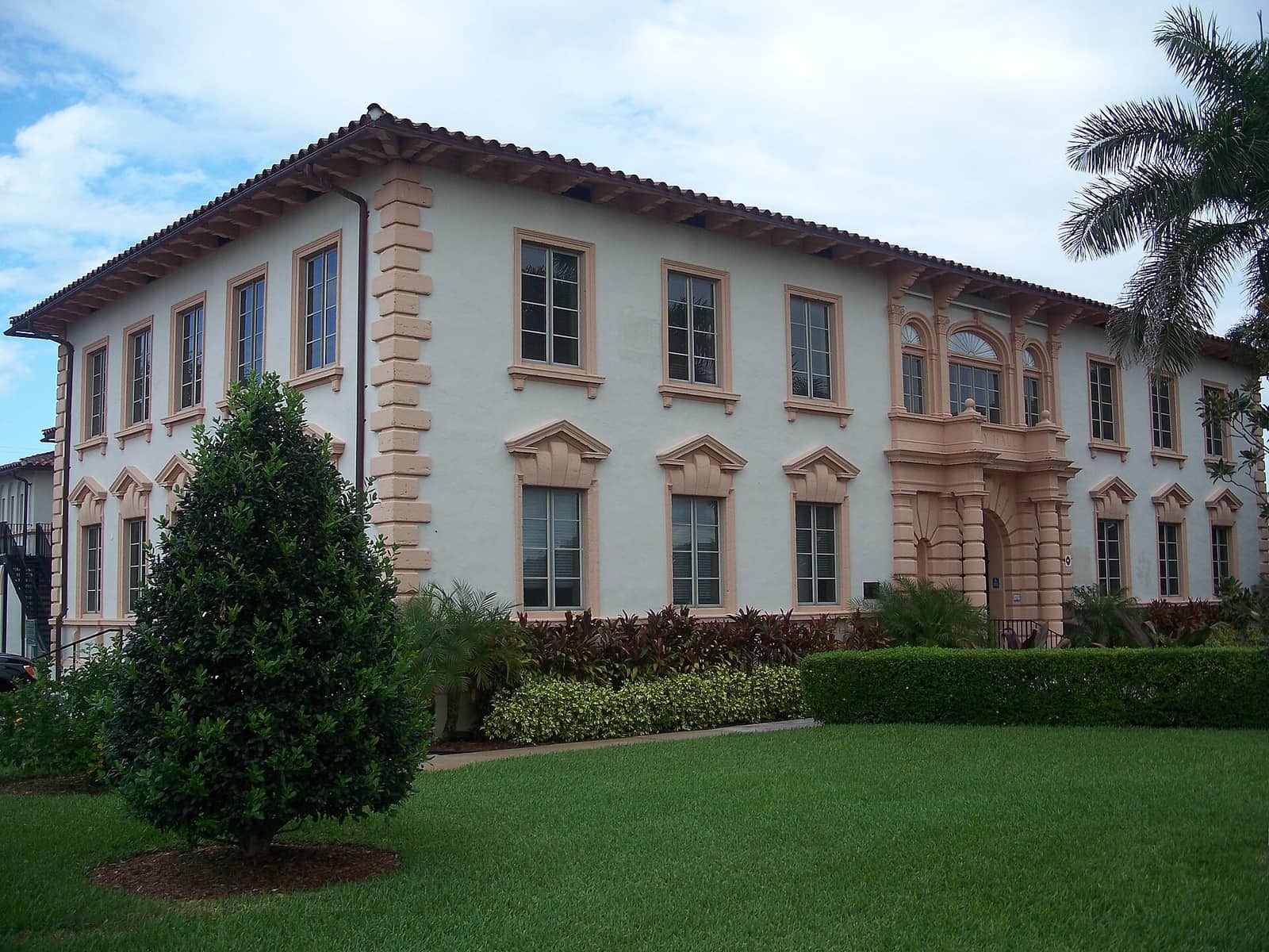 Two-story cream stucco civic building with red tile roof, arched windows, and palm trees beside a lawn.