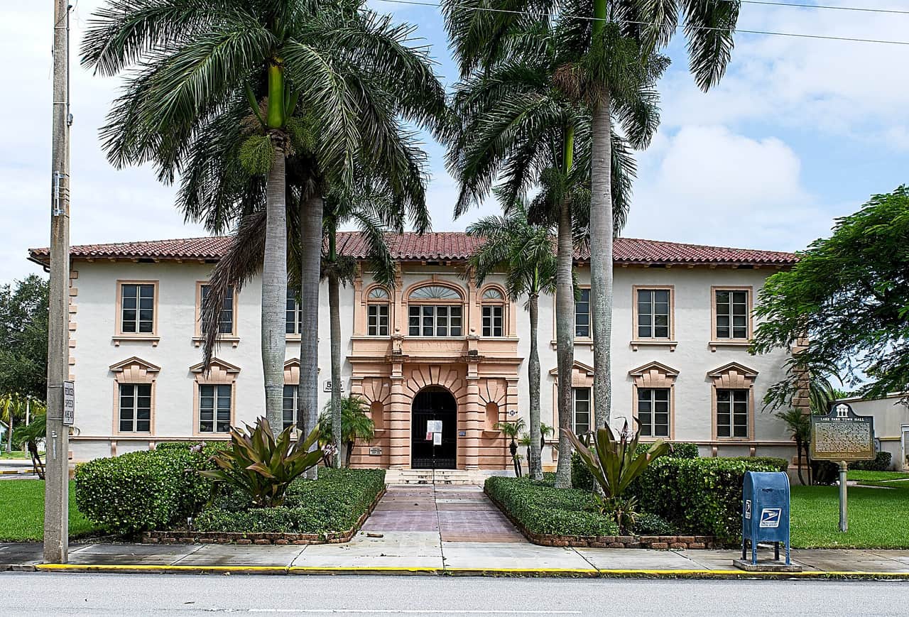Cream stucco civic building with palm trees, a blue mailbox and red tile roof beside a street.