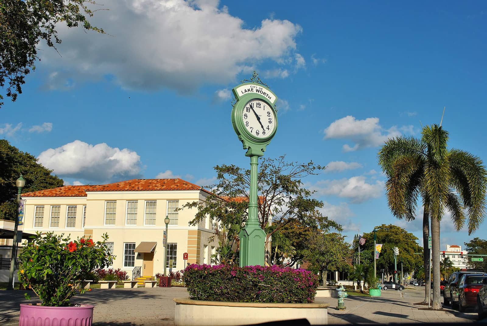 Green street clock with a Lake Worth sign stands beside a cream building and palm trees.