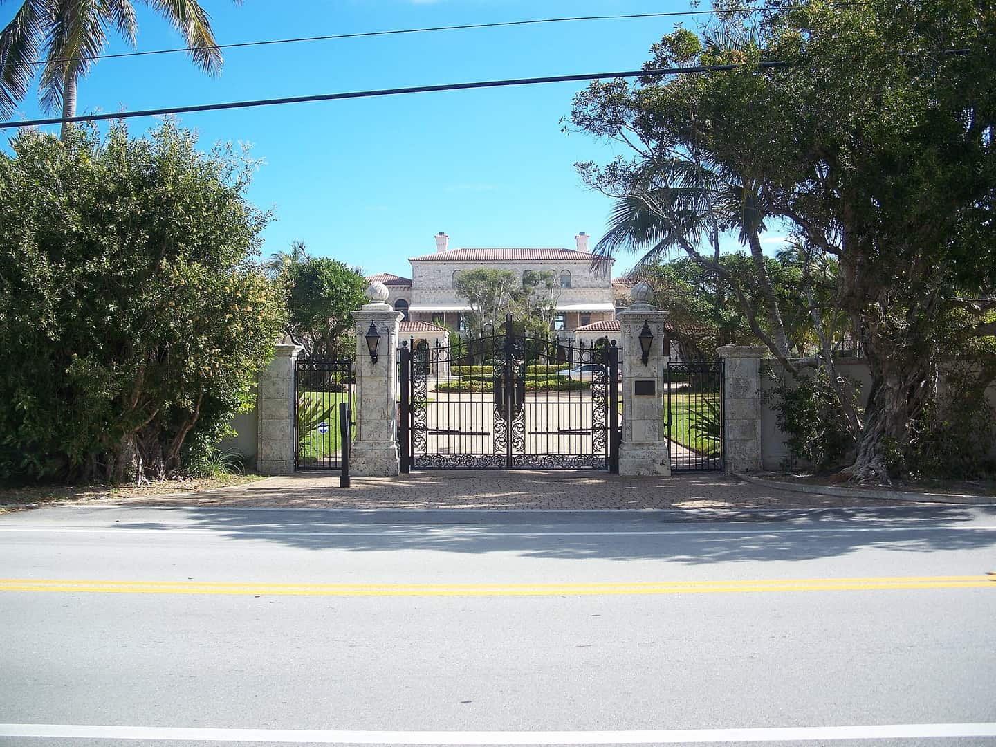 Ornate iron gates and stone pillars at the entrance to a large estate, with a house behind.