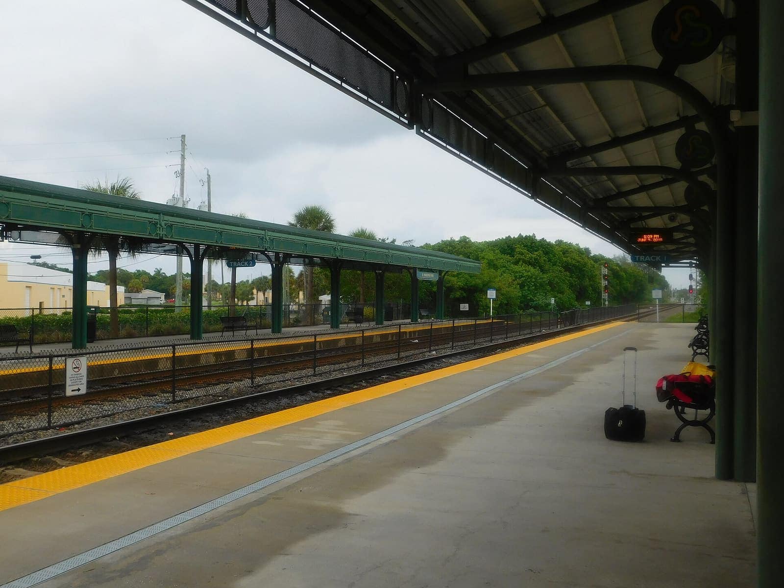 Empty commuter rail platform under a green-roofed canopy with a suitcase and bench.
