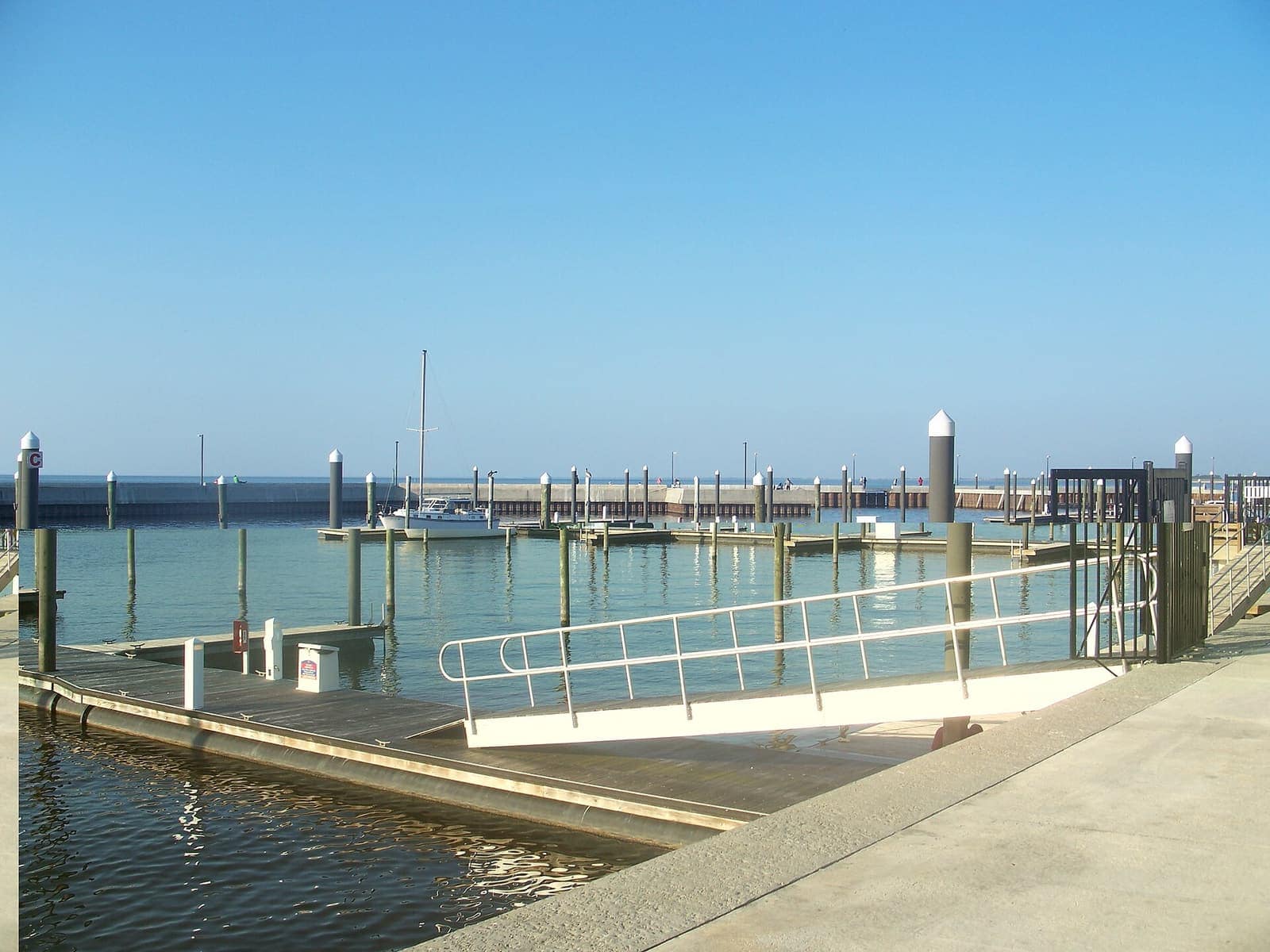 Boat marina with floating docks, moored boats and a railed gangway on calm water under clear blue sky.