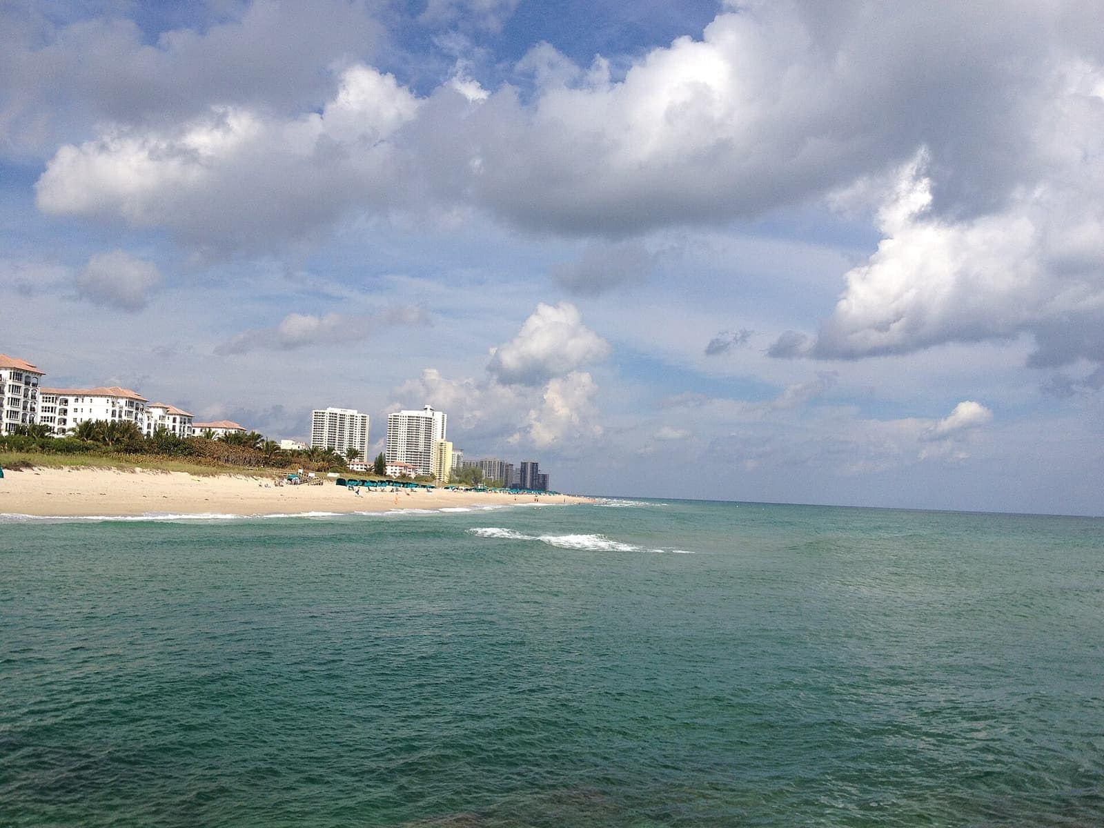 Sandy beach lined with white beachfront condo towers under cumulus clouds above turquoise ocean water.