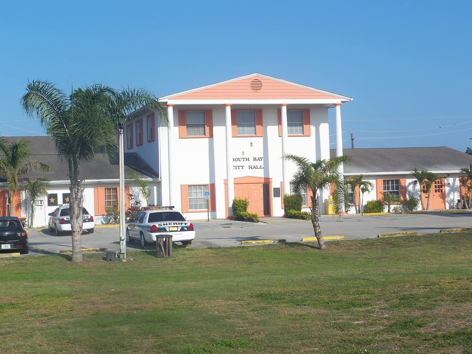 White city hall with peach trim, parked cars and a police vehicle in a daytime lot.