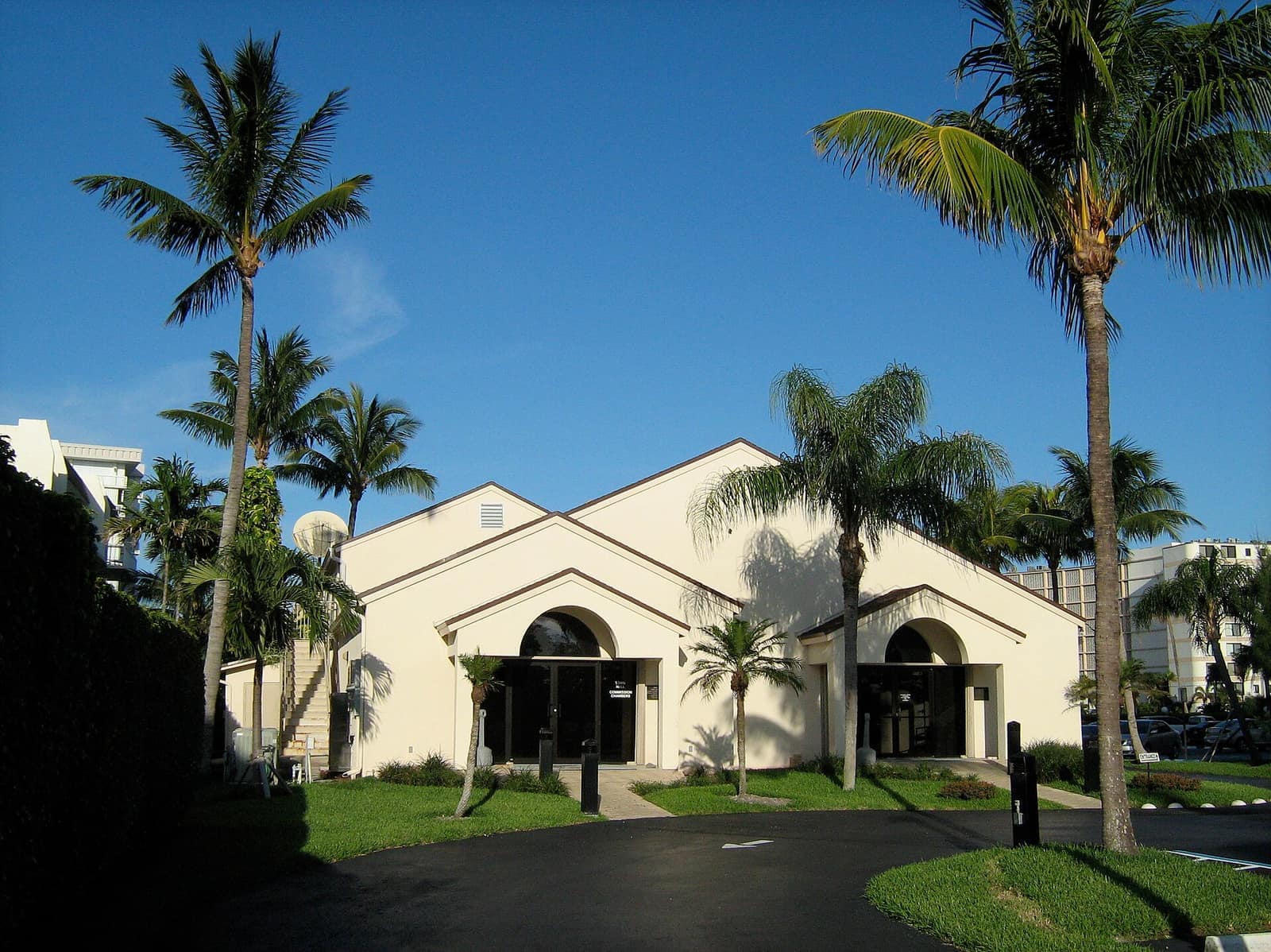 Cream-colored municipal building with arched entrances, palm trees and blue sky in daylight.