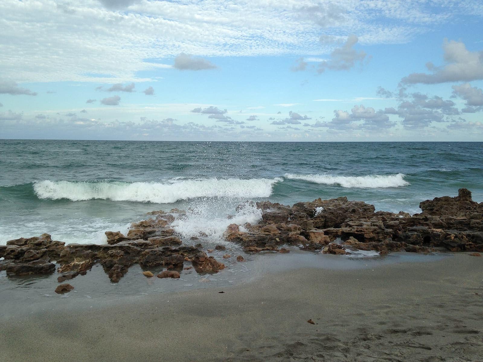 Waves breaking over craggy limestone rocks on a sandy beach under blue sky.