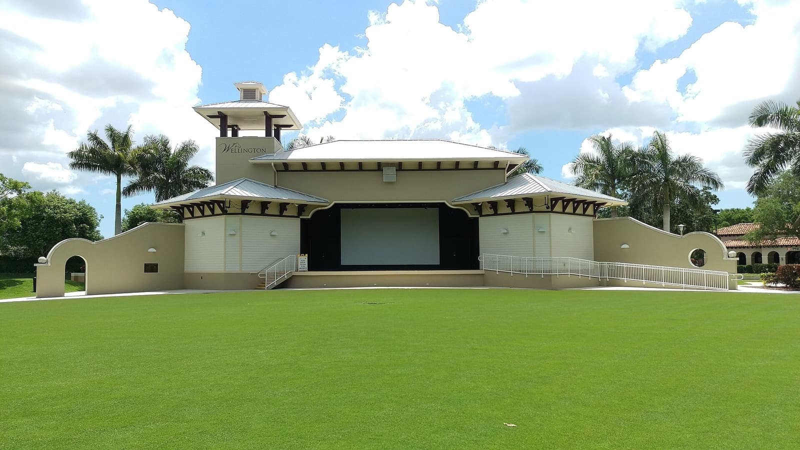 Outdoor amphitheater stage with a green lawn, palm trees and blue sky with clouds.