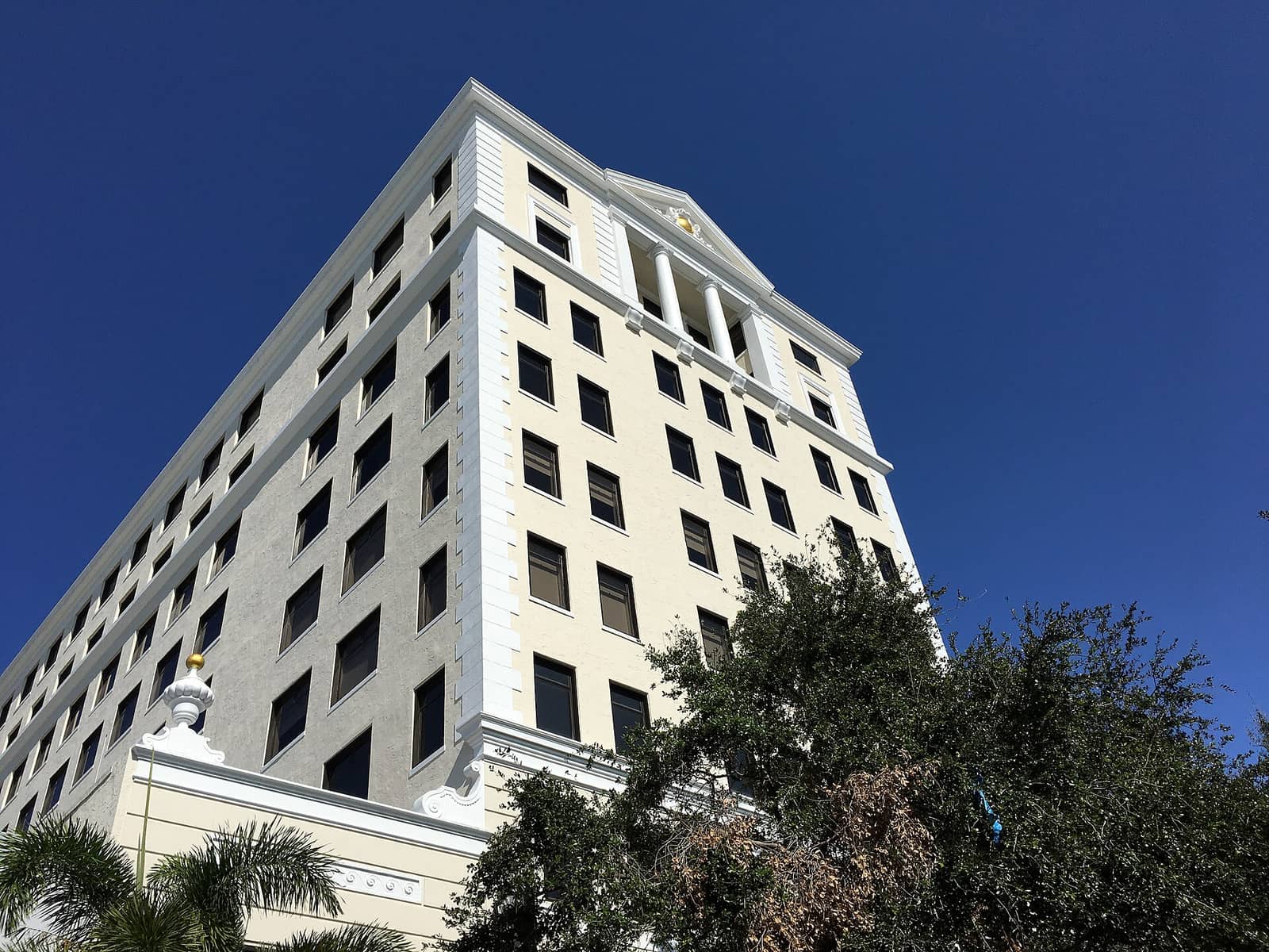 Tall white historic tower with classical top rising against a deep blue sky above green trees.