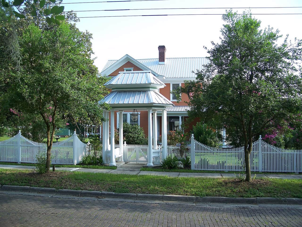 Two-story brick house with white picket fencing and a metal-roofed gazebo-style porch under shade trees.