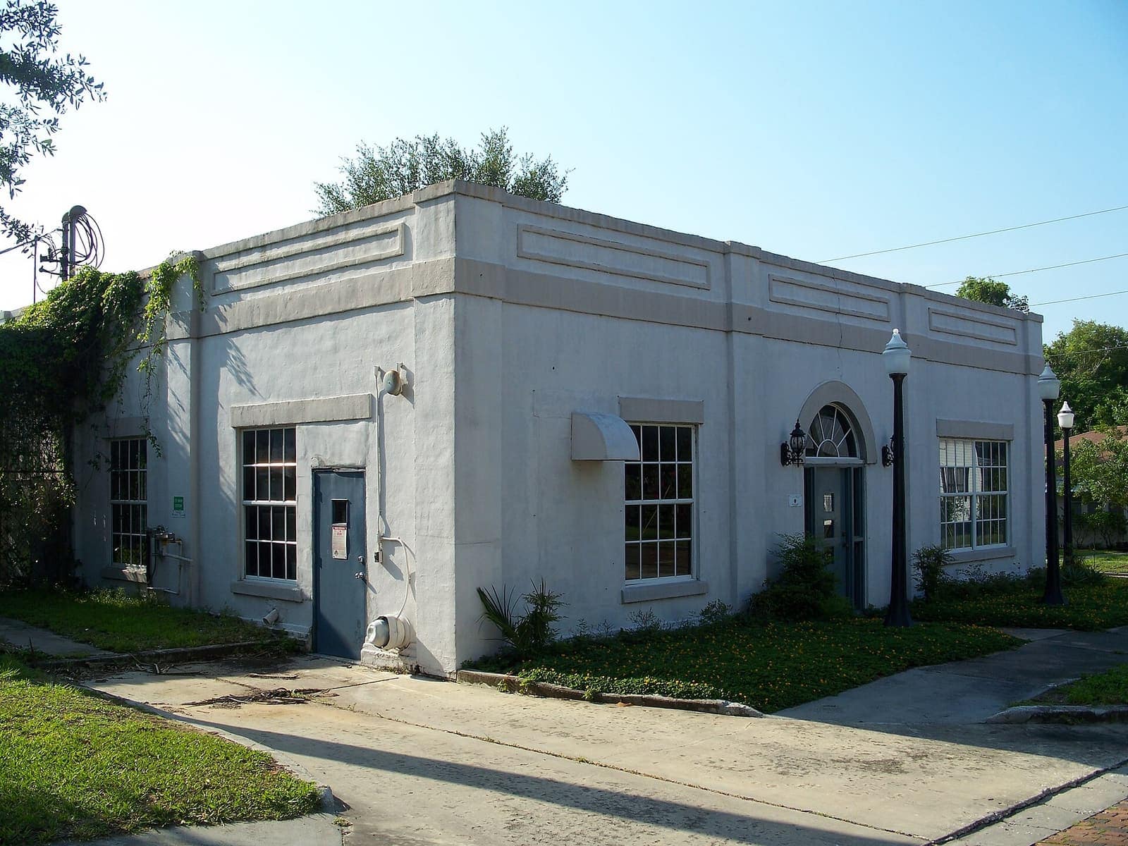Single-storey white stucco building with arched entry door and street lamps beside a concrete walkway.