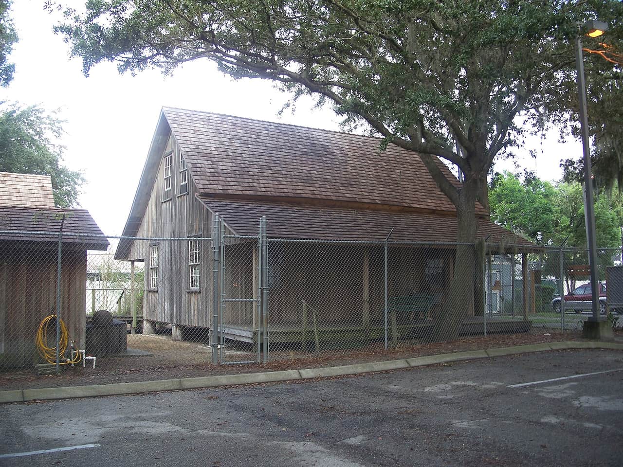 Weathered wooden historic house with a shingled roof behind chain-link fencing beside a large oak tree and an empty parking lot.