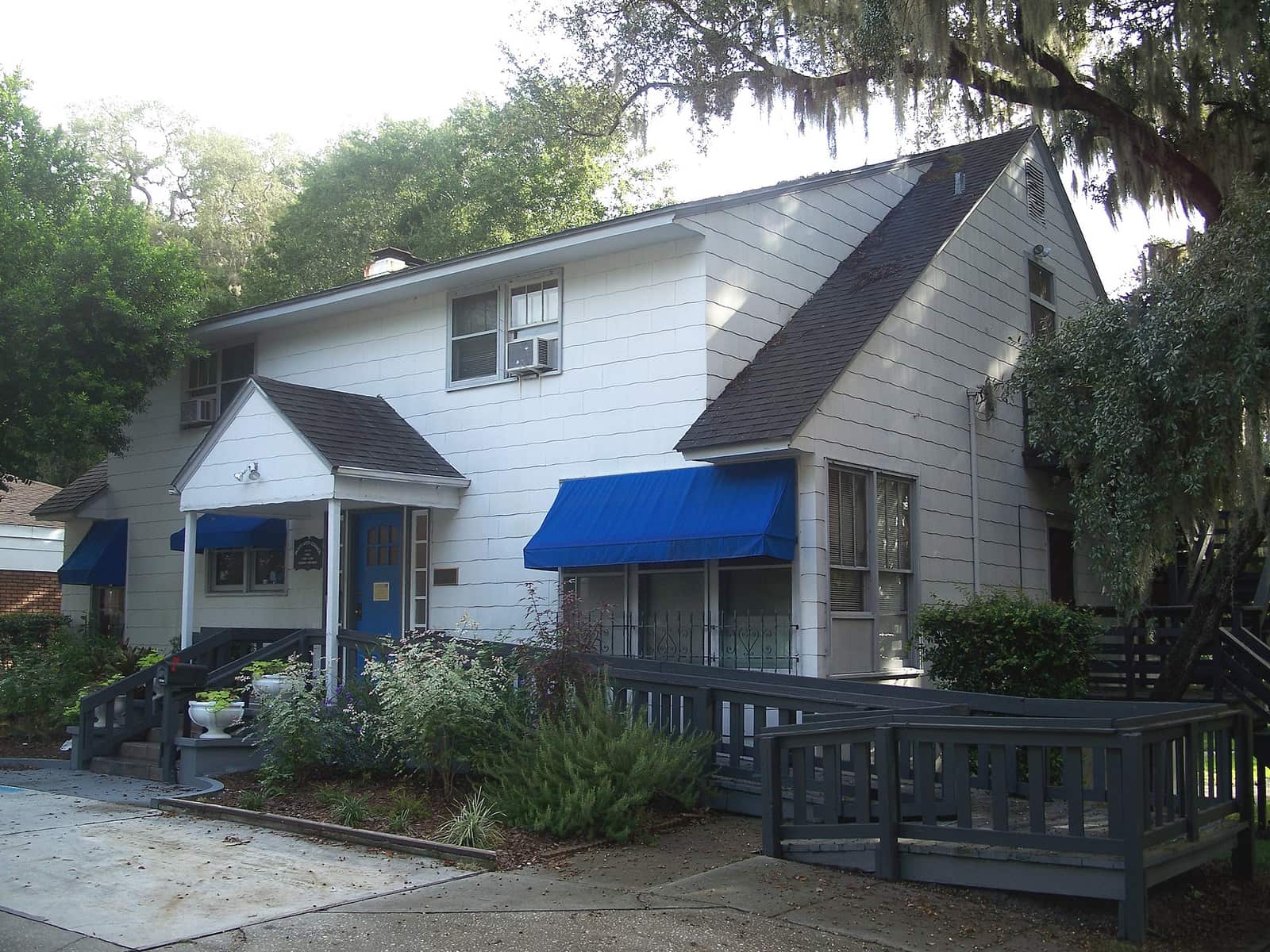 White wood-sided cottage with blue awnings, a wooden deck, and shrubs under a large tree with hanging moss.