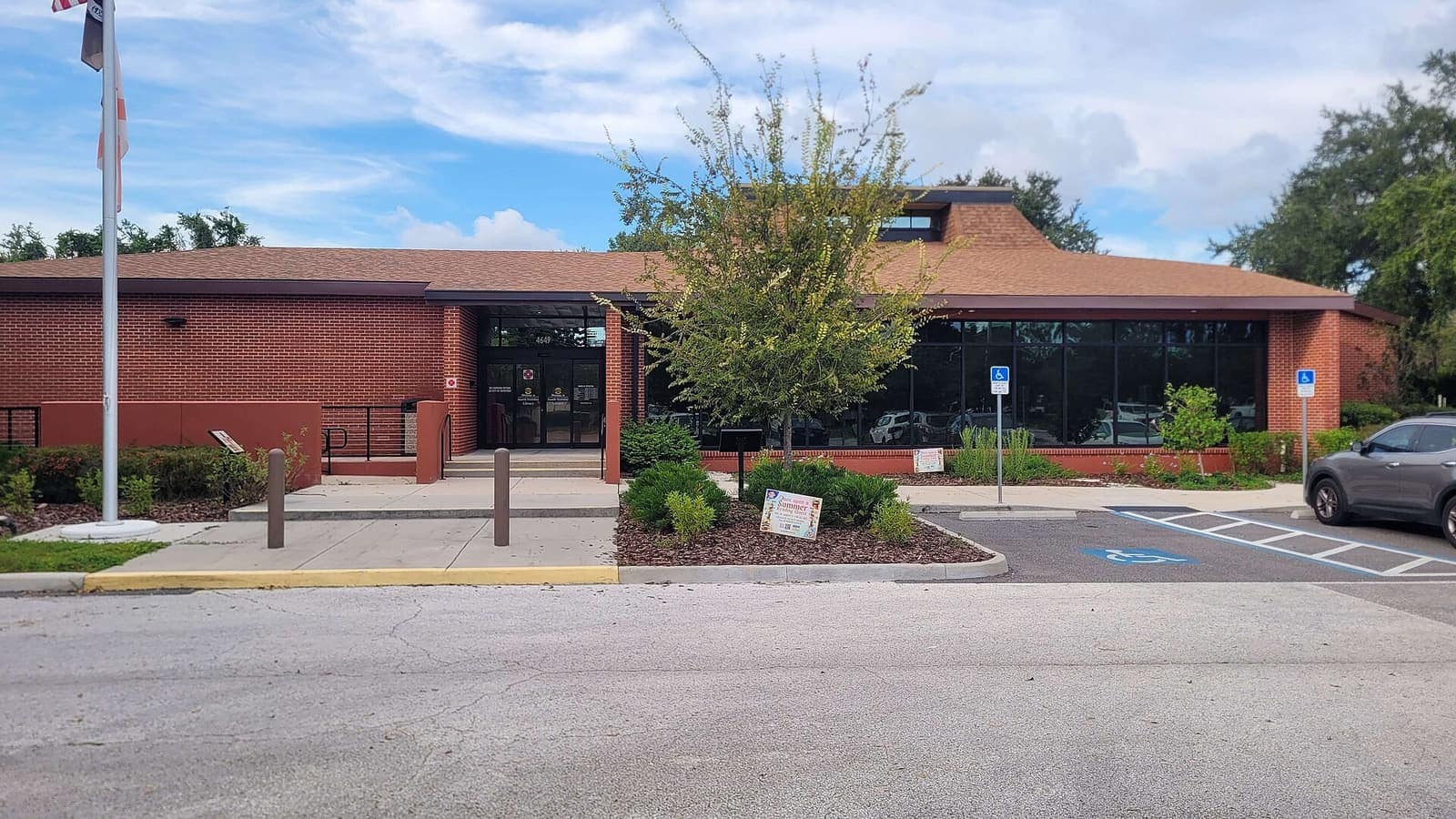 Single-storey brick library building with glass entrance, flagpole, landscaped beds and a parking lot under blue sky.