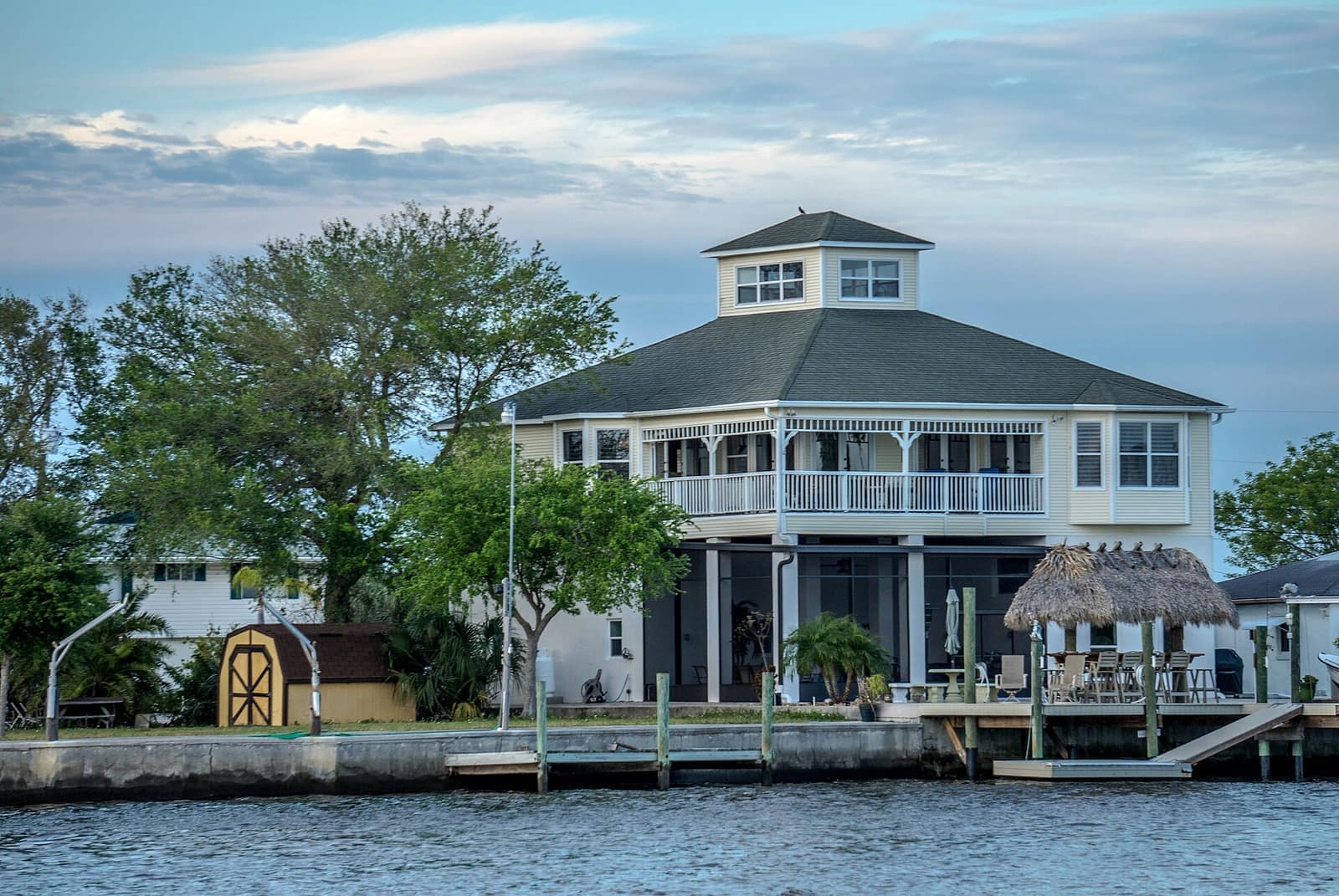 Two-story waterfront home with a covered balcony and tiki hut beside a canal dock at dusk.