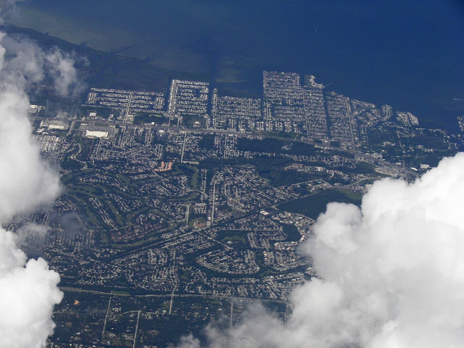 Aerial view of a dense residential neighborhood with canals and a waterfront edge, partly framed by clouds.