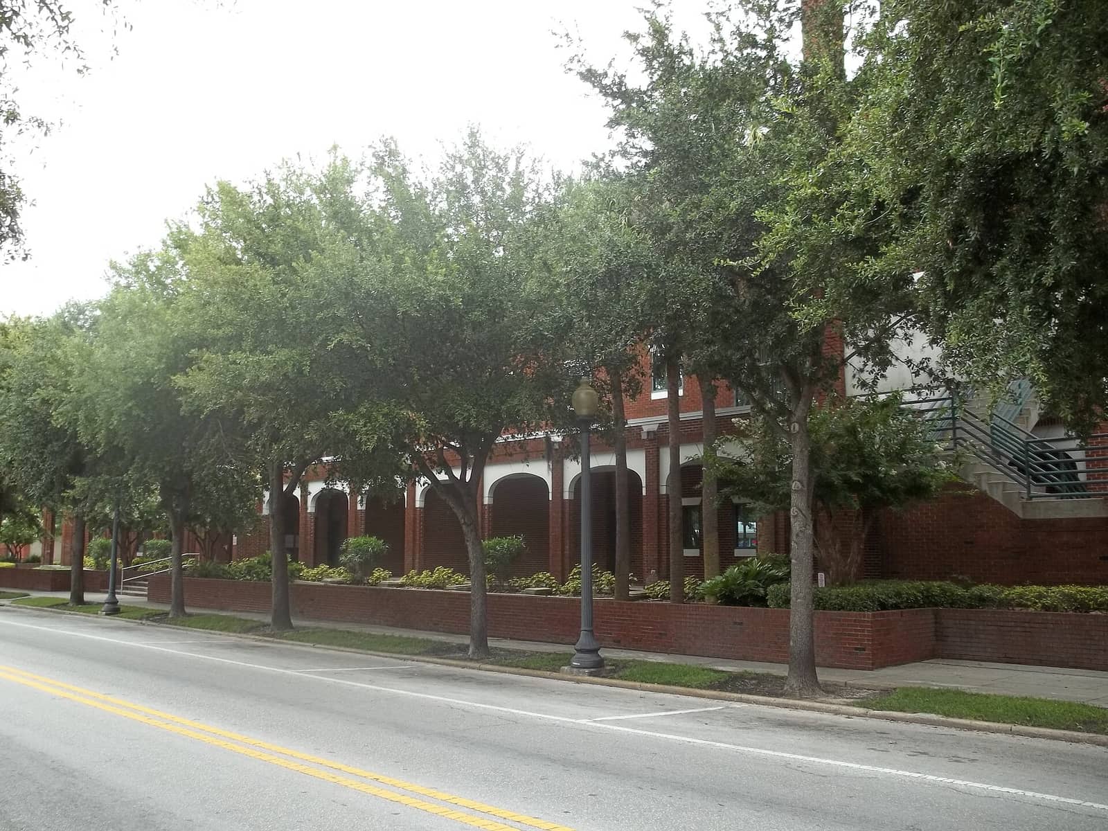 Brick city hall building with arched openings and trees lining a street on an overcast day.