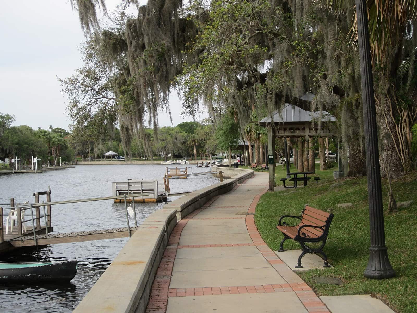 Paved riverside walkway with a bench, gazebo and moss-draped trees beside a calm waterway.