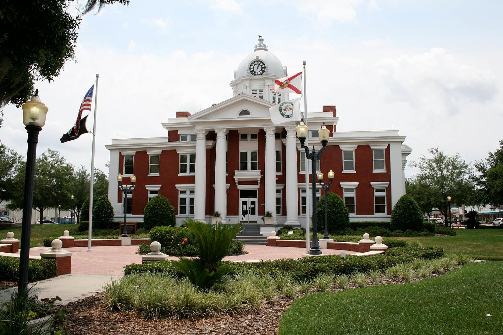 Red brick courthouse with white columns and a clock tower behind lampposts, flags and trimmed shrubs.