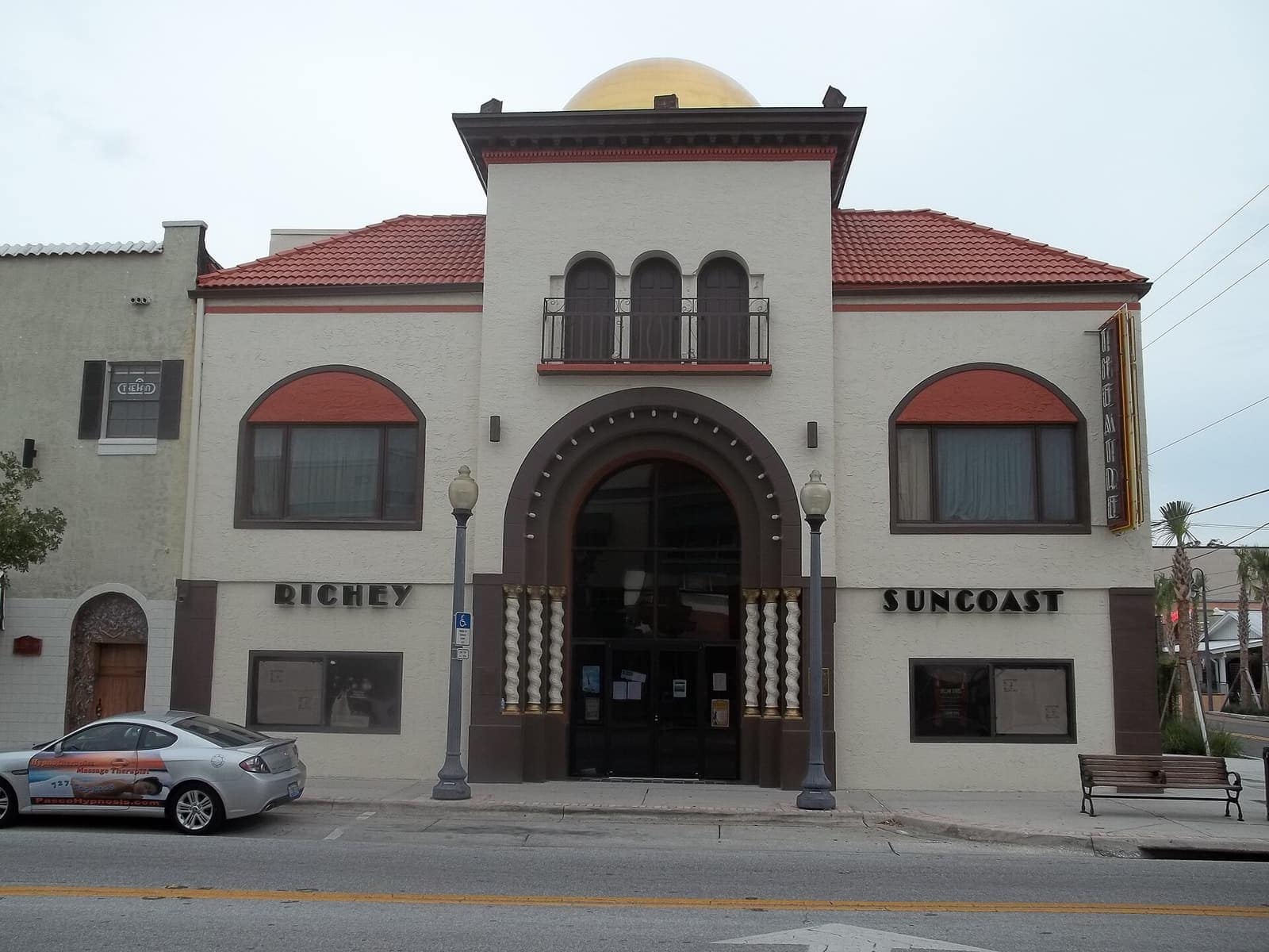 Cream stucco theater facade with red tile roof, arched entrance, and a gold dome under overcast sky.