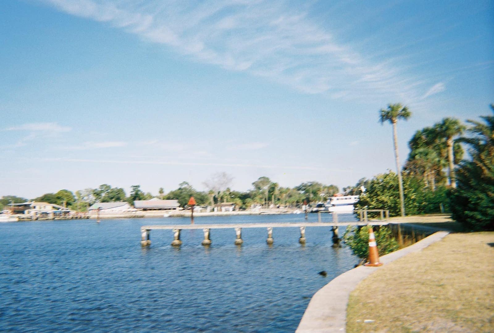 Calm river with a wooden dock on pilings, palm trees, and a white boat under a blue sky.